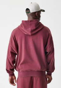 Bordo kapućast pulover iz mehkih materialov, s sproščenim krojem, rebrastimi manšetami in robom. Zadnja stran prikazuje minimalističen dizajn.