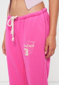 Pink sweatpants with a drawstring waist, featuring embroidered branding and number "3" on the side. Soft material and relaxed fit.