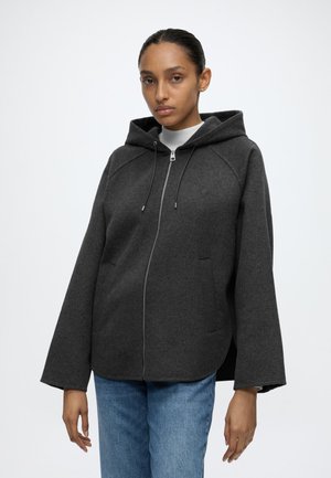 Donna che indossa una giacca con cappuccio e zip di colore grigio scuro con maniche larghe e jeans blu, in piedi davanti a uno sfondo bianco uniforme.