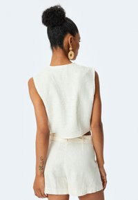 Blusa senza maniche color crema con tessuto testurizzato, abbinata a pantaloni corti a vita alta dello stesso colore. Orecchini a cerchio e un tatuaggio visibile sul braccio.