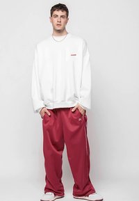 Muž stojí so založenými rukami v vreckách, oblečený v bielom oversize svetri a voľných bordových nohaviciach so postrannými pruhmi a bielych teniskách.