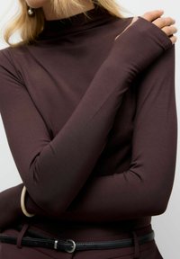 Maglietta rossa a maniche lunghe con collo alto e design aderente. Materiale morbido e liscio con fori per i pollici alle cuciture delle maniche.