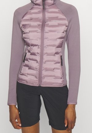 Veste matelassée violette zippée à manches longues et short noir portée par une personne debout, mains détendues le long du corps.
