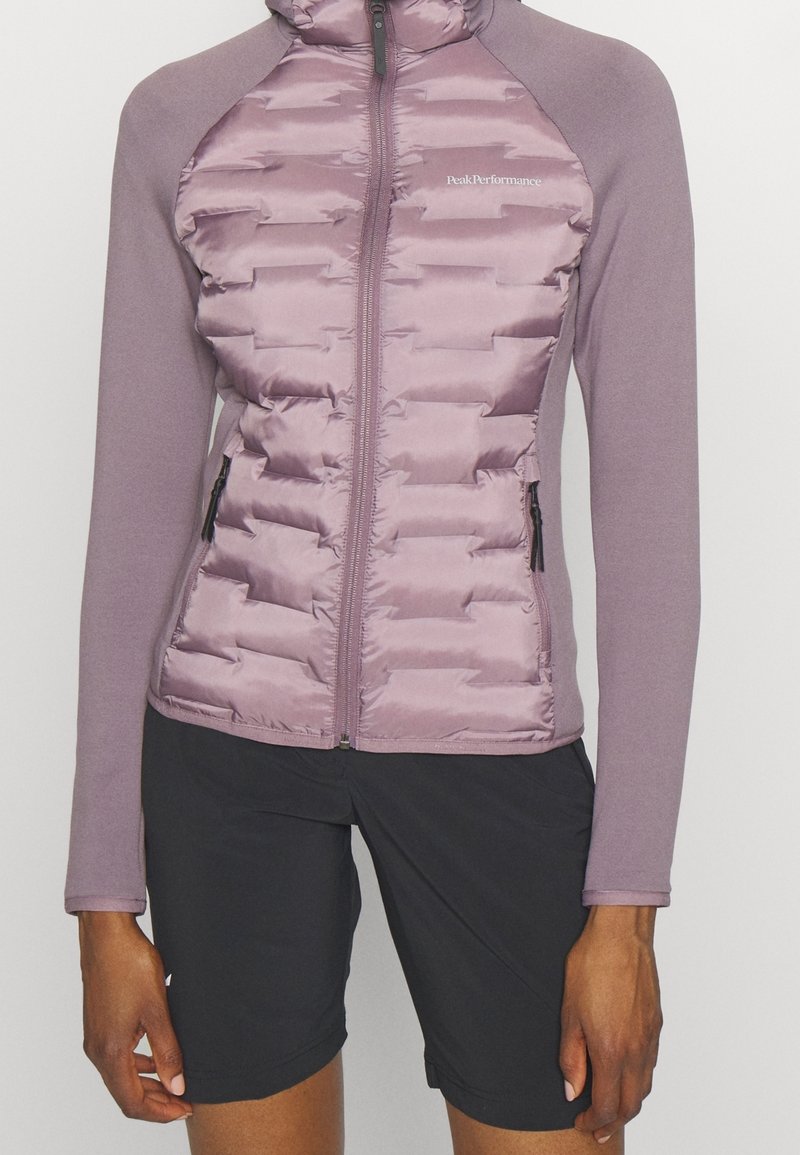 Veste matelassée violette zippée à manches longues et short noir portée par une personne debout, mains détendues le long du corps.