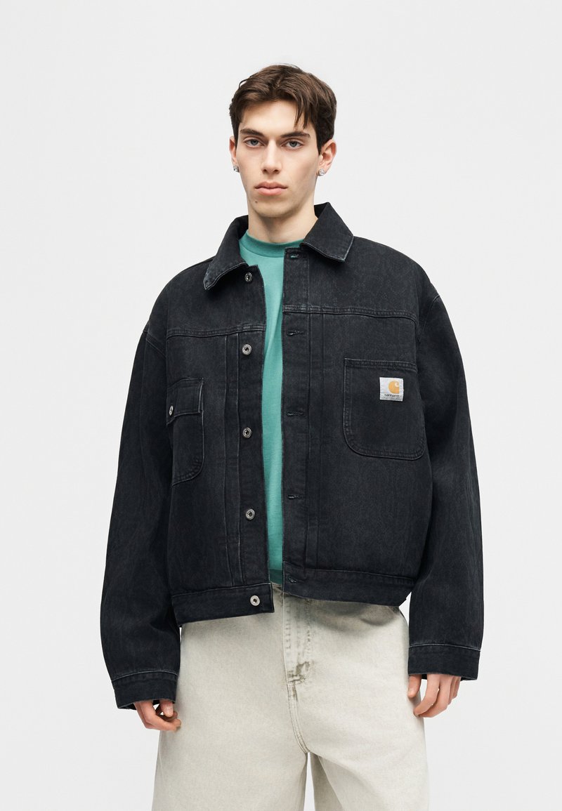 Jonge man met kort bruin haar, gekleed in een zwart oversized spijkerjack over een groen shirt en lichtbeige broek, staand tegen een effen achtergrond.