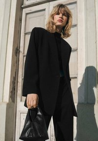 Schwarzer, maßgeschneiderter Blazer aus Stoff mit strukturierter Silhouette. Kombiniert mit einem schwarzen Top, eleganten schwarzen Hosen und einer kleinen Lederhandtasche.