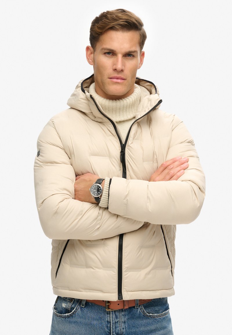 Beige pufferjack met een capuchon, gestructureerd oppervlak, zwarte rits en zijzakken. Draag het over een crèmekleurige coltrui, met een aansluitend ontwerp.