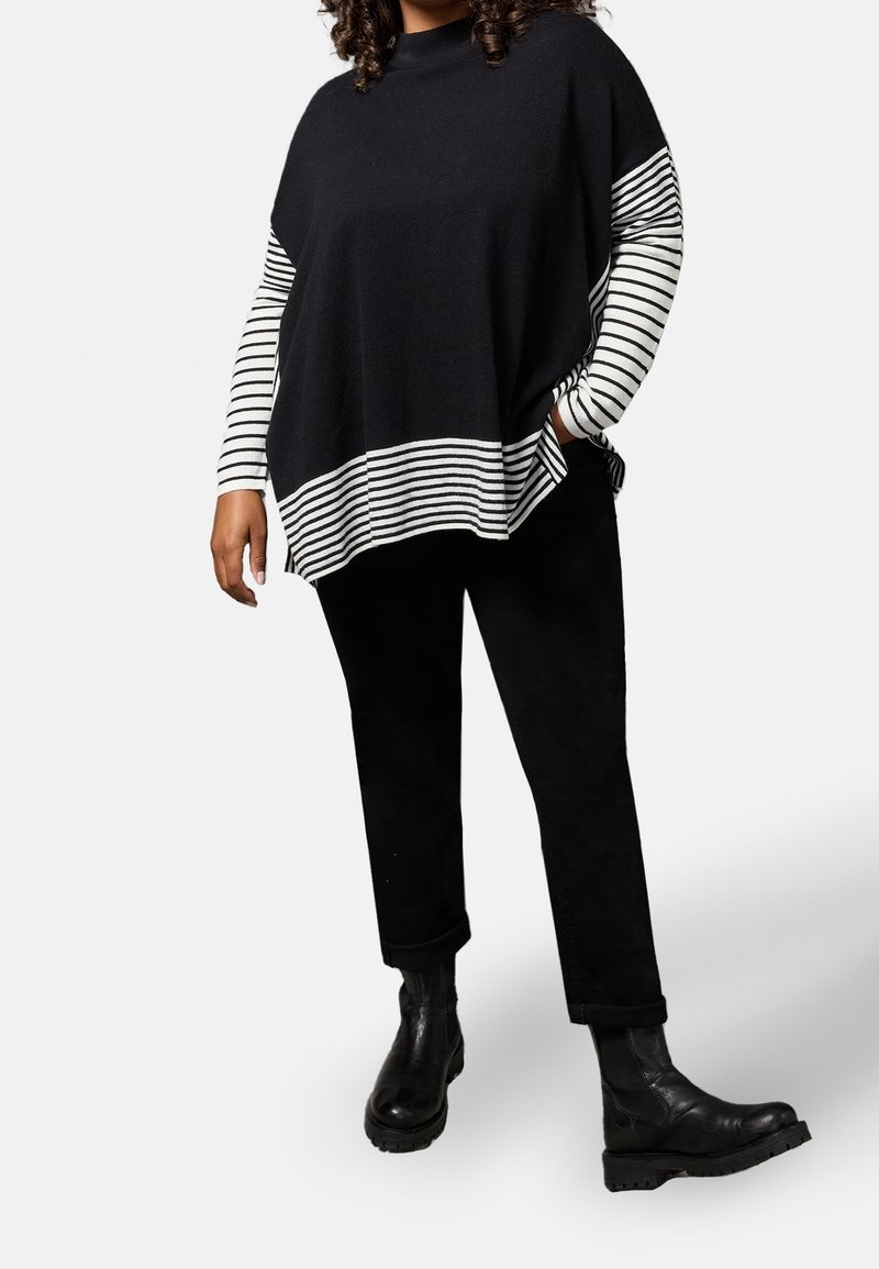 Personne portant un pantalon noir, des bottes en cuir noir et un pull noir avec des manches et un ourlet rayés blanc et noir, debout sur fond blanc.