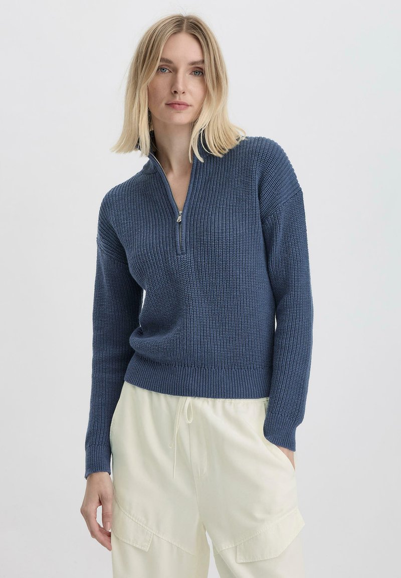 Blauwe pullover trui met een halve ritssluiting, geribbelde textuur en aangesloten manchetten, gecombineerd met crèmekleurige wijde broek.