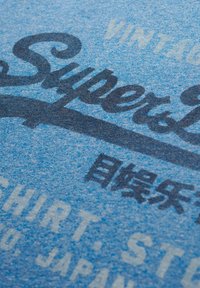 Blå stof T-shirt med vintage-stil mørkeblå tryk. Teksten inkluderer "Super" og "T-shirt," samt japanske tegn og en struktureret overflade.