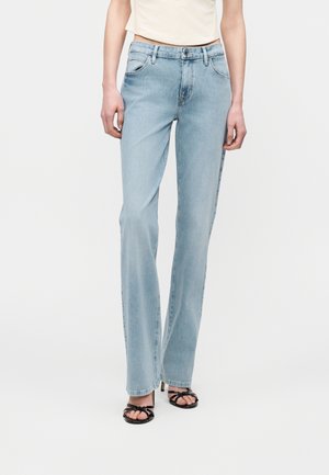 Žena oblečená v svetlomodrých džínsach s rovným strihom a prednými vreckami, s čiernymi sandálmi na vysokom podpätku s remienkami, stojaca pred jednoduchým pozadím.