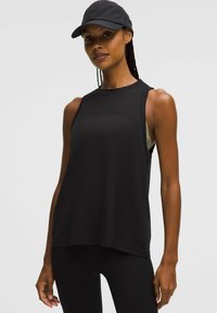 Fit vrouw in een zwarte mouwloze sporttop, zwarte leggings en een zwarte pet, staande tegen een effen lichte achtergrond.