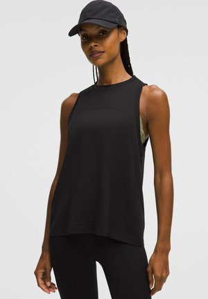 Fit woman wearing black sleeveless workout top, black leggings, and black cap, standing against plain light background.