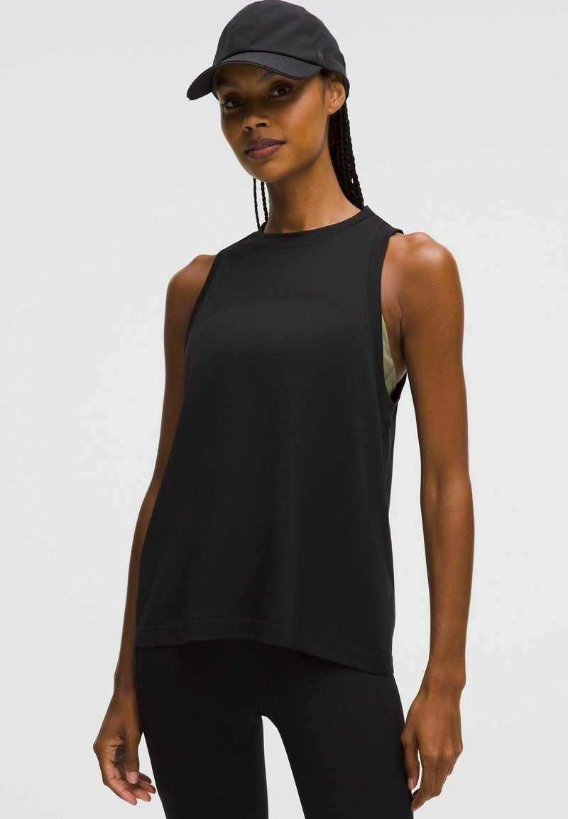 Fit vrouw in een zwarte mouwloze sporttop, zwarte leggings en een zwarte pet, staande tegen een effen lichte achtergrond.