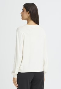 Pull en tricot blanc à coupe décontractée, manches longues et poignets côtelés. Présente une texture subtile et un col rond, relevé à l'arrière.