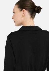 Crna majica s ovratnikom, glatke teksture i minimalnim detaljima, prikazana sa stražnje strane. Zlatne naušnice su vidljive na modelu.