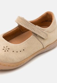 Zapato Mary Jane de ante beige con una correa, diseño floral recortado en la punta y suela de goma color marrón. Superficie texturizada con forro suave.