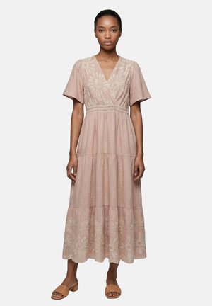 Donna che indossa un abito midi rosa chiaro con maniche corte e ricamo floreale traforato, abbinato a sandali slip-on intrecciati color cuoio.