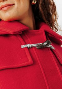 Manteau en laine rouge avec un col haut, présentant une fermeture à crochets et des accents en corde grise. Texture lisse avec des poches latérales et une coupe ajustée.