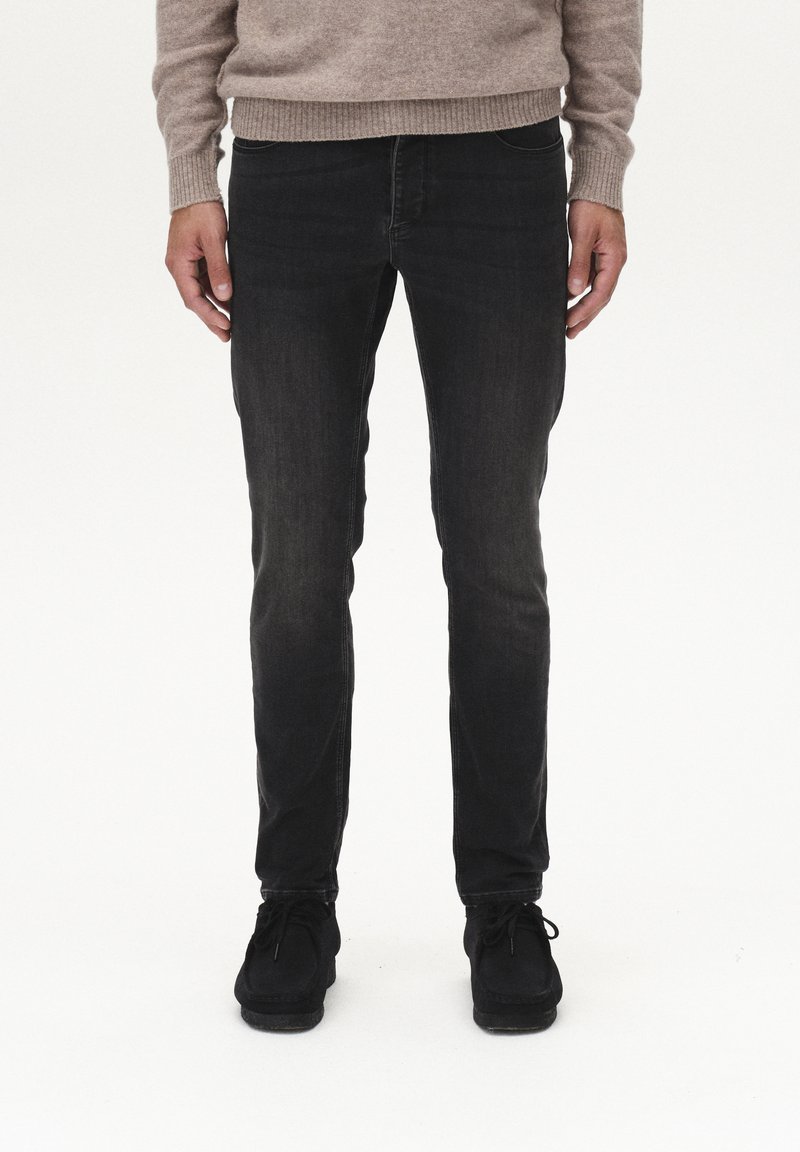 Schwarze Jeans mit einer slim fit Passform und leichter Verwaschung, kombiniert mit einem beigen Strickpullover. Die Schuhe sind aus schwarzem Wildleder.