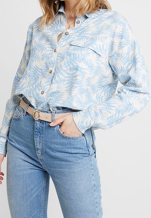 Kobieta w jasnoniebieskiej koszuli w wzory, włożonej do wysokich niebieskich dżinsów z beżowym paskiem, z ręką w kieszeni.