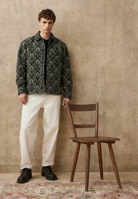 Schwarz-weiße Jacke mit Muster und Reißverschluss, weiße Hose und schwarze Schuhe. Neben einem Holzstuhl auf einem strukturierten beigen Hintergrund.