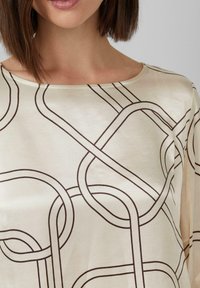 Szatynowa bluzka w kremowym kolorze z geometrycznym brązowym wzorem, z dekoltem w łódkę i luźnym krojem, podkreślająca gładką teksturę.