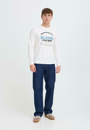 Langärmliges weißes Baumwoll-T-Shirt mit grafischem Text, kombiniert mit dunkelblauen Jeans. Legere Schuhe runden das Outfit ab.