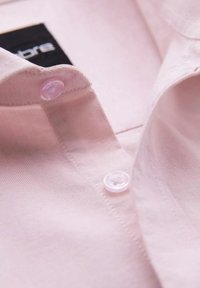 Lichtroze overhemd met kraag van een gemêleerde stof en twee doorzichtige knopen bij de hals. Bevat een zwart label dat binnenin de kraag is genaaid.