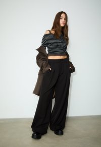 Zwarte wijde broek gecombineerd met een grijze gestreepte cropped top. Een bruine jas hangt over de arm. Gladde stof, losse pasvorm, minimalistisch ontwerp.