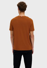 T-shirt marrone a maniche corte con collo rotondo, realizzato in tessuto morbido. Presenta un piccolo accento tricolore sul retro del collo. Design semplice e aderente.