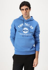 Blauer Kapuzenpullover aus weichem Material mit Kängurutasche, verstellbarem Kapuzenzug und weißem Drucklogo sowie Text auf der Vorderseite. Kombiniert mit schwarzen Hosen.