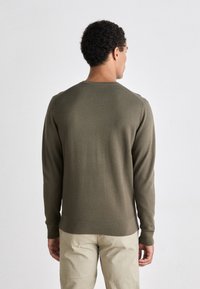 Maglione verde oliva con texture, maniche lunghe e polsini a costine, dotato di scollatura rotonda e fondo aderente. Indossato con pantaloni beige chiaro.