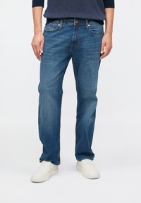 Pantalones vaqueros de mezclilla en azul medio con corte recto, diseño de cinco bolsillos y un sutil desgaste; combinados con zapatos blancos sin cordones.