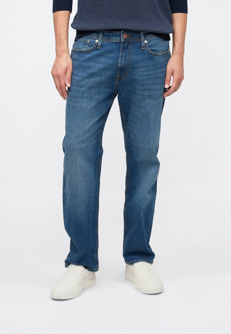 Pantalones vaqueros de mezclilla en azul medio con corte recto, diseño de cinco bolsillos y un sutil desgaste; combinados con zapatos blancos sin cordones.