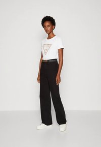 Femeie în picioare, purtând un tricou alb cu logo-ul Guess, pantaloni negri cu talie înaltă și lățime largă cu o curea neagră, și adidași albi cu platformă.