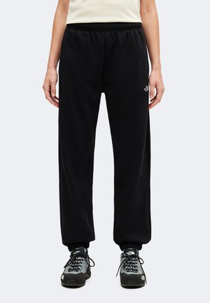 Personne portant un pantalon en polaire noir avec des poignets élastiques et des chaussures de randonnée grises, debout devant un fond clair uni.