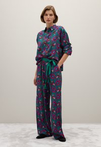 Kleurrijke pyjama set met een groene basis en rode en blauwe paisleypatronen, een knoopsluiting shirt, broek met elastische tailleband en een bijpassende groene riem.