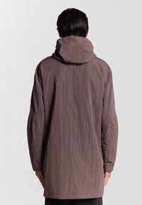 Lång, lätt jacka i en mauvefärg med huva, texturerat tyg och sänkta axlar. Bakvy visar en minimalistisk design.
