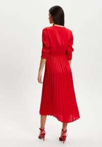 Red pleated dress with long sleeves, cinched waist, flowing silhouette, and a high-low hem. Paired with red high-heeled shoes.