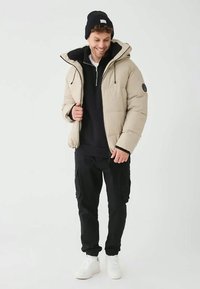 Uomo che indossa un giubbotto trapuntato beige, un berretto nero, pantaloni cargo neri e sneakers bianche, sorridente e con lo sguardo rivolto verso il basso su uno sfondo semplice.