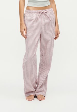 Femme portant un pantalon ample rayé rose et blanc avec une taille à cordon, debout pieds nus sur un fond uni.
