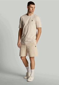 Beige cotton t-shirt with short sleeves and a small logo on the chest, paired with matching beige shorts, worn with white socks and sneakers.