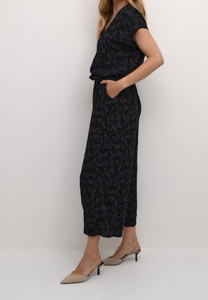 Combinaison noire à motifs floraux avec un décolleté en v, des manches courtes, une taille élastiquée, des poches latérales et une coupe décontractée ; associée à des mules à talons beiges.