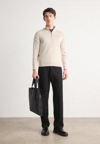 Suéter de punto beige con cuello de cremallera, combinado con pantalones negros y un bolso de piel negro, con tela texturizada y un diseño sencillo.