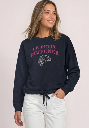 Femme portant un sweat-shirt bleu marine avec un texte rose "Le Petit Déjeuner" et une illustration de croissant, assorti à un pantalon blanc, souriante et regardant vers la droite.