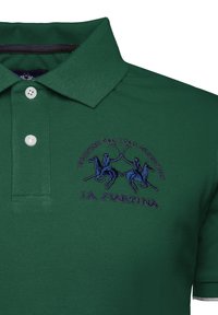 Polo shirt verde realizzato in tessuto strutturato, con un logo ricamato viola che raffigura due cavalieri e tre bottoni sul colletto.