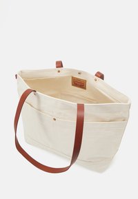 Krämfärgad canvas toteväska med bruna läderremmar, två ytterfickor och en innerfoder i bomull som har en varumärkesetikett.
