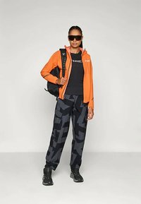 Jachetă ușoară portocalie peste un tricou negru, asociată cu pantaloni gri cu un model și adidași negri. Accesorii includ ochelari de soare și un rucsac negru.