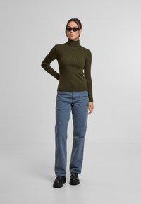 Ein olivgrüner, gerippter Rollkragenpullover, kombiniert mit hellblauen, schmal geschnittenen Jeans und schwarzen Schuhen mit dickem Sohlenprofil, vor einem einfarbigen Hintergrund.